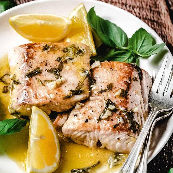 Grilled fish fillets with lemon slices and fresh basil, served on a white plate with herbs and garlic
