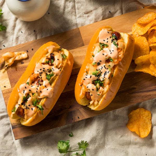 Gourmet hot dogs with creamy sauce, herbs, and sesame seeds served on wooden board with potato chips